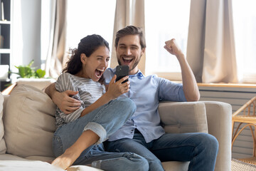 Overjoyed surprised young couple looking at phone screen, reading message with good news, excited woman and man celebrating success, online lottery win, showing yes gesture, sitting on couch