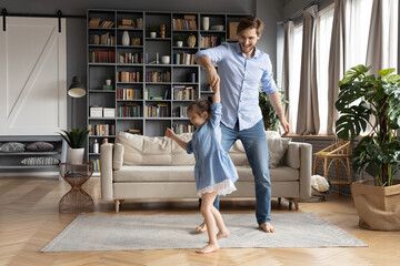 Happy father dancing with adorable little daughter wearing dress, holding hands, moving to favorite music in living room, family having fun, smiling dad and cute girl enjoying leisure time together - Powered by Adobe