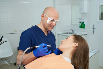 Fototapeta premium A bald smiling middle-aged dentist in a medical face shield started to treat woman's teeth with a dental drill. A doctor and his female patient during a dental procedure in a dentist's office.