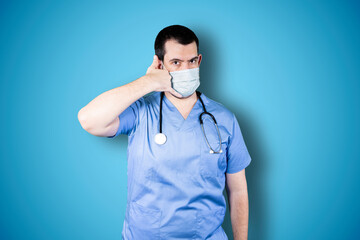 Handsome bearded doctor with mask making phone gesture over blue studio background