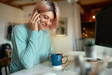 Fashionable young female copywriter with pink hair and facial piercing sitting at her workplace in front of desktop computer, holding cup, drinking coffee and having phone conversation, smiling