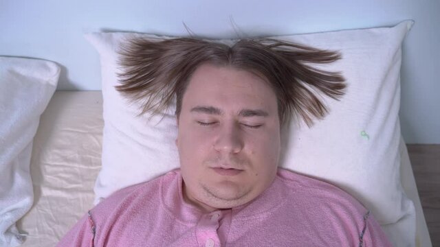 Young unshaven plump man with ridiculous long bangs divided into the two sides of the head, sleeps in his bed, slightly snoring. Wears funny pink pajamas. Humor concept of comical hairstyle.