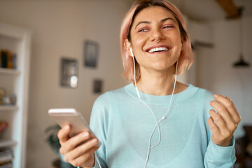 Fashionable young woman with pink hair enjoying online communication, talking to friend on video conference call using earbuds and wifi on smart phone. Happy pretty girl listening to music at home