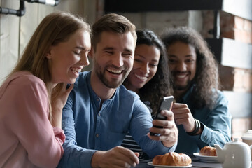 Cheerful diverse multi racial millennial people sitting at table in cozy cafe eat pastries drink tea watch funny entertainment video use smartphone device, best friends having fun spends time together