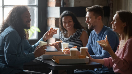 Mixed-race caucasian friends sit at table in cafe eat pizza drink coffee having funny talk. Sharing ordered pizza slices spend Friday in pizzeria yummy fast food. Racial equality, friendship concept