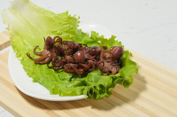 Cooked octopuses are placed on a lettuce leaf on a plate. Bamboo cutting Board and texture table as background.