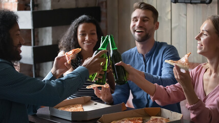 Multi racial excited friends eating pizza hold bottles with alcohol beverage clinking with beer, celebrate success, life event in cafe, relaxing together in public place. Friendship, lifestyle concept