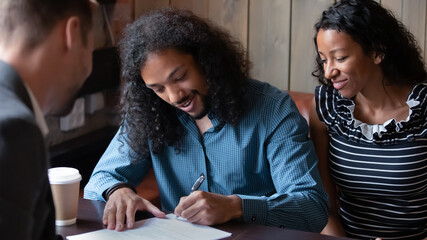 African ethnicity 35s couple sign contract document closing deal with real estate agent sit in public place cafe. Happy spouses put signature on agreement taking obligations with banker getting loan