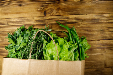 Paper bag with green onion, rosemary, lettuce leaves and parsley on wooden table. Top view. Healthy food and grocery shopping concept