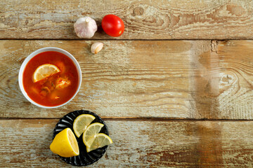 A dish with thai tom yam soup on a wooden table and garlic and lemon nearby.