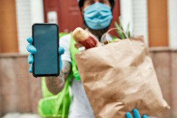 Bearded delivery man wearing mask and gloves due to the emergence of the Covid19 virus, showing smartphone, holding grocery bag while delivering food