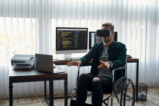 VR at work. Young male web developer in a wheelchair wearing virtual reality glasses while working at his workplace in modern office. High Tech