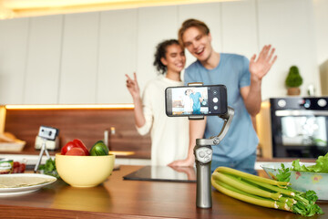 Obraz premium Happy couple, vegetarians smiling, recording a video on smartphone while going to prepare a healthy meal in the kitchen together. Vegetarianism, healthy food, blog concept