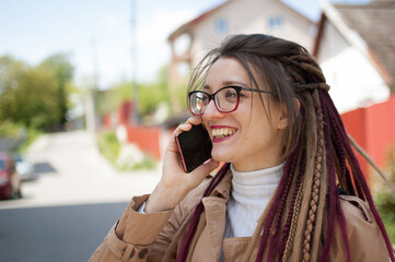 Modern young business woman with long dreadlocks and eyeglasses is talking with the client by mobile phone standing at the street near the buildings