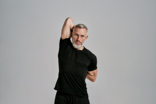 Middle aged muscular man in black sportswear looking at camera while warming up his body before workout in studio over grey background