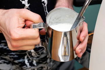 barista whips milk in a pitcher with a steam of a coffee machine