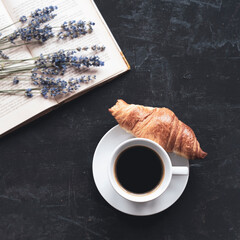 Cup of coffee with croissant, book and lavender bouquet on black table. Top view, flat lay