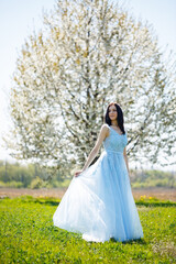 Fototapeta premium Portrait of a girl in a light summer blue long dress adorned in her hair against a blossoming tree.
