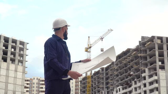 Portrait of a successful young engineer, architect, builder, businessman, wearing a white helmet, in a shirt, holding a project in his hand, a skyscraper background and a construction site