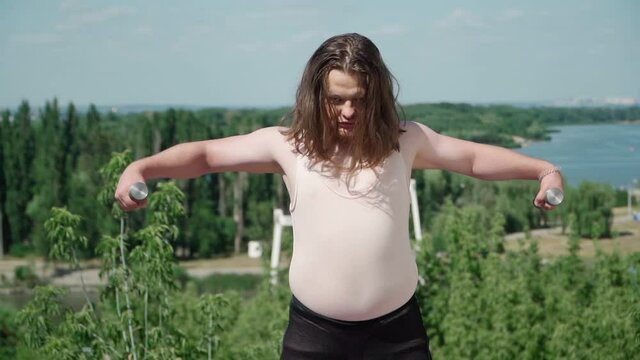 Playful Fat Man with Long Hair Does Gymnastics for Weight Loss outdoors. Funny Overweight Guy Depicting a Girl Engaged In Fitness with Dumbbells In A Park. Slow motion