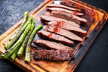 Traditional barbecue sliced dry aged wagyu flank steak offered with green asparagus and chili powder as close-up on a rustic burnt wooden cutting board
