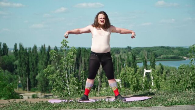 Playful Fat Man with Long Hair in Pink T-shirt and Tight Leggings Does Gymnastics for Weight Loss outdoors. Funny Overweight Guy Depicting Girl Engaged In Fitness with Dumbbells In Park. Slow motion