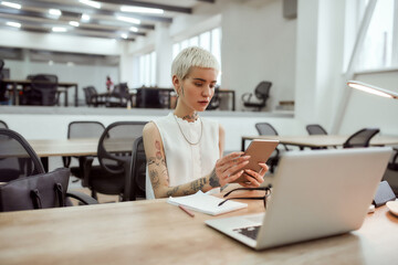 Full concentration. Portrait of young stylish blonde tattooed business woman with short haircut using touchpad while sitting at her working place in the modern office. Digital technologies at work