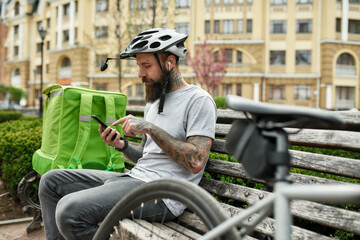 Just try it. Brutal bearded delivery man in helmet with thermo bag or backpack checking order using smartphone, sitting on the bench outdoors. Courier, delivery service concept