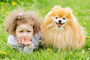 Pensive little beautiful girl lies on the grass with dog. baby hugs red, ginger pomeranian spitz outdoors. small doggy guards a child. favorite pet. female loves animal. waiting for parents