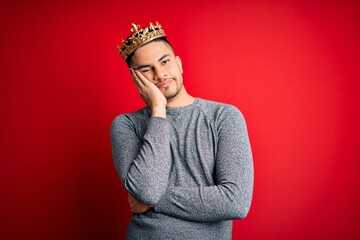 Young handsome man wearing golden crown of prince over isolated red background thinking looking tired and bored with depression problems with crossed arms.