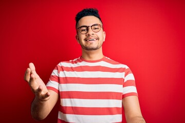 Young handsome man wearing casual striped t-shirt and glasses over isolated red background smiling cheerful with open arms as friendly welcome, positive and confident greetings