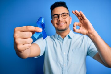 Naklejka premium Young handsome man holding blue cancer ribbon standing over isolated background doing ok sign with fingers, excellent symbol
