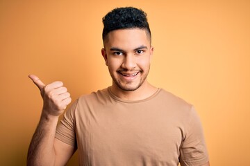 Young handsome man wearing casual t-shirt standing over isolated yellow background smiling with happy face looking and pointing to the side with thumb up.