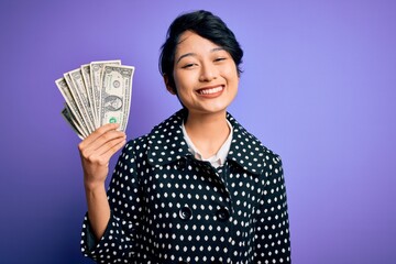 Young beautiful asian girl holding bunch of dollars banknotes over purple background with a happy face standing and smiling with a confident smile showing teeth