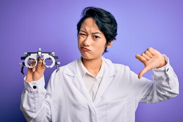 Beautiful asian optical girl wearing coat holding optometry glasses over purple background with angry face, negative sign showing dislike with thumbs down, rejection concept