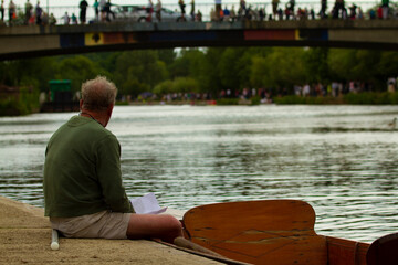 An elderly man is sitting on concrete ground by the river with his feet in a wooden boat. He is watching a rowing event and checking his notes. Other spectators are seen across the river and on bridge