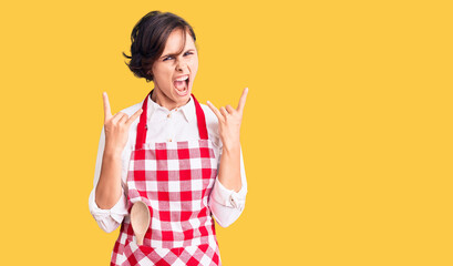 Beautiful young woman with short hair wearing professional cook apron shouting with crazy expression doing rock symbol with hands up. music star. heavy concept.