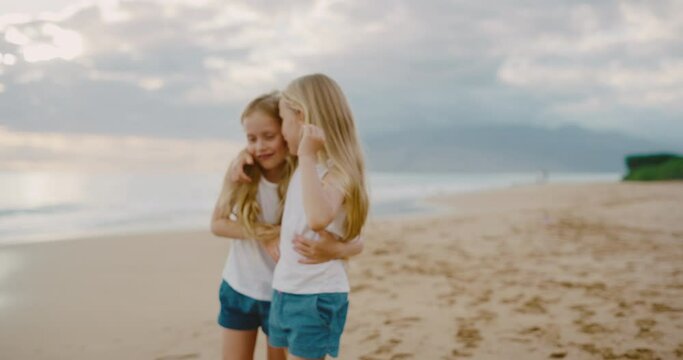 Happy young twin girls walking down the beach playing at sunset, summer family lifestyle