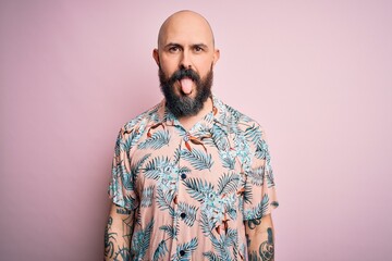 Obraz premium Handsome bald man with beard and tattoo wearing casual floral shirt over pink background sticking tongue out happy with funny expression. Emotion concept.