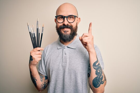 Handsome bald artist man with beard and tattoo painting using painter brushes surprised with an idea or question pointing finger with happy face, number one