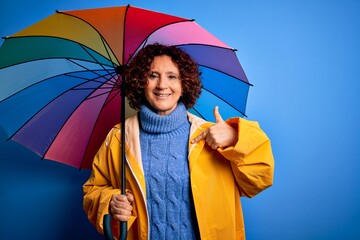 Middle age curly hair woman wearing rain coat holding colorful umbrella over blue background with...