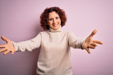 Middle age beautiful curly hair woman wearing casual turtleneck sweater over pink background looking at the camera smiling with open arms for hug. Cheerful expression embracing happiness.