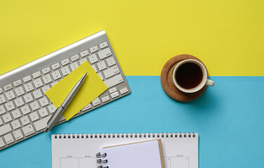 Flatlay of coffee, wireless keyboard, notepad, calendar and yellow paper on yellow and blue tabletop