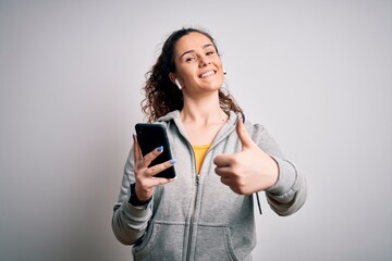 Beautiful sportswoman with curly hair listening to music using smartphone and earphones happy with big smile doing ok sign, thumb up with fingers, excellent sign