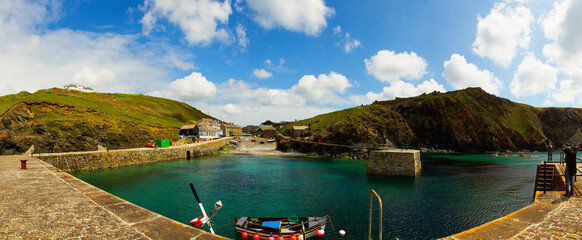A scenic little fishing harbor in a small cove in Cornwall. A few houses, beautiful skyline, wharf quay, boat ramp and small fishing boats are seen. Some people are seen on the dock