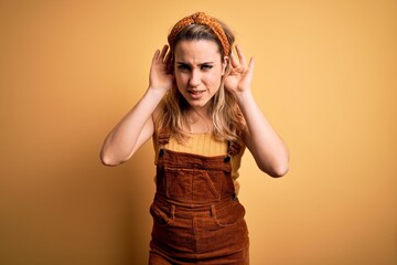 Young beautiful blonde woman wearing overalls and diadem standing over yellow background Trying to hear both hands on ear gesture, curious for gossip. Hearing problem, deaf