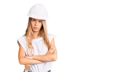 Beautiful caucasian woman with blonde hair wearing hardhat and painter clothes skeptic and nervous, disapproving expression on face with crossed arms. negative person.