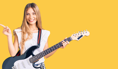 Fototapeta premium Beautiful caucasian woman with blonde hair playing electric guitar smiling happy pointing with hand and finger to the side