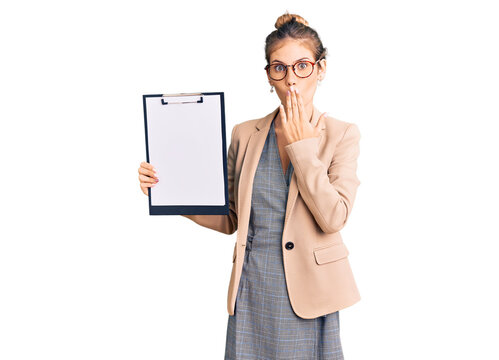 Beautiful caucasian woman with blonde hair holding clipboard with blank space covering mouth with hand, shocked and afraid for mistake. surprised expression