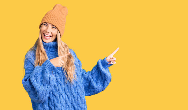 Beautiful caucasian woman with blonde hair wearing wool sweater and winter hat smiling and looking at the camera pointing with two hands and fingers to the side.